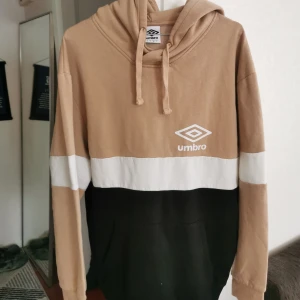 Beige, vit och svart hoodie från Umbro - Snygg hoodie från Umbro i beige, vitt och svart med stor logga framtill. Tröjan har huva med snörning och en klassisk känguruficka. Perfekt för chill dagar eller när du vill ha en sportig vibe.