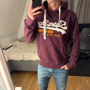 Superdry hoodie - Säljer en vinröd hoodie från Superdry som är sjukt snygg och väldigt bra skick för att vara såhär eftertraktad, passa på denna är såld inom kort!