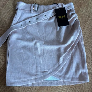 Rosa omlottkjol från Dennis Maglic - Snygg rosa kortkjol från Dennis Maglic i mjukt material med omlott-design och bälte med silverfärgat spänne. Kjolen har dekorativa nitar på bältet och en lätt draperad framsida som ger en trendig look. Kjolen ser ut att vara mer beige, men är mer ljusrosa/rosa. 