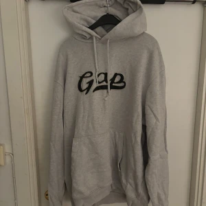 Grå GAP hoodie - Säljer en grå hoodie från GAP i storlek L. Tröjan har en broderad GAP-logga i svart framtill, stor huva med snörning och en klassisk känguruficka. Tillverkad i mjuk bomulls- och polyestermix, perfekt för chill dagar.