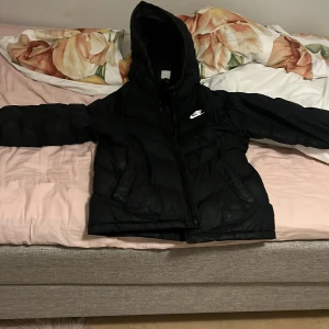 Svart pufferjacka från Nike - Säljer en svart pufferjacka från Nike med huva och dragkedja framtill. Jackan har en klassisk Nike-logga på bröstet och två fickor på sidorna. Perfekt för kyliga dagar och enkel att matcha med det mesta. Priset kan diskuteras 