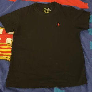 Svart t-shirt från Polo Ralph Lauren - Svart t-shirt från Polo Ralph Lauren med klassisk röd broderad logga på bröstet. T-shirten har rund halsringning och korta ärmar, perfekt för en clean och stilren look. Tillverkad i mjuk bomull som känns skön mot huden.