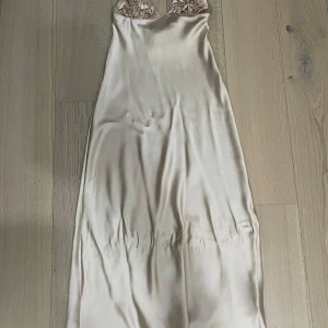 Champagnefärgad långklänning i satin - Elegant långklänning i skimrande champagnefärgad satin med smala axelband och dekorativa blommor vid bysten. Klänningen har en slank siluett och faller snyggt längs kroppen, perfekt för speciella tillfällen.