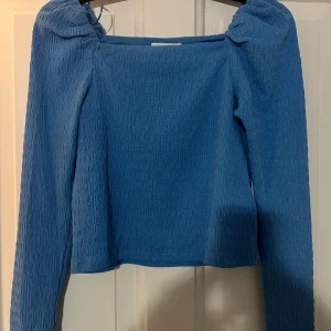Blå långärmad topp från H&M - Säljer en blå långärmad topp från H&M med fyrkantig ringning och lätt puffade axlar. Toppen har en ribbad struktur och är tillverkad i ett stretchigt material som sitter snyggt på kroppen. Perfekt för att lyfta din outfit med en pop av färg.
