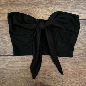 Svart bandeau tubtopp med knyt - Snygg svart bandeau tubtopp med knyt framtill. Toppen är ribbad och har en croppad passform som sitter tight runt kroppen. Perfekt för att skapa en trendig look med hög midja eller kjol.