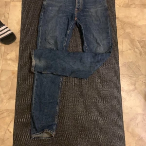 Blå jeans från Nudie Jeans, W29 L32 - Säljer ett par klassiska blå jeans från Nudie Jeans med orange sömmar och snygga bakfickor med vågdetalj. Modellen har normal passform och raka ben. Tillverkade i slitstarkt jeanstyg av bomull. Perfekta för dig som gillar stilrena och tidlösa jeans.