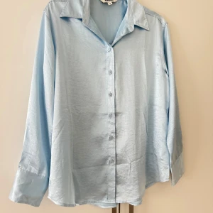 Ljusblå silkesliknande skjorta KappAhl - Snygg ljusblå skjorta från KappAhl med silkesliknande finish. Klassisk krage, knappar hela vägen och långa ärmar med breda manschetter. Perfekt för dig som gillar stilrena och fräscha plagg med lite extra glans.