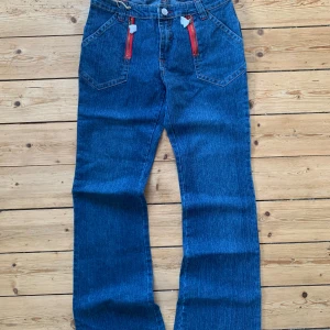 Blå jeans från Ginger med röda dragkedjor W30 L32 - Snygga blå jeans från Ginger med raka ben och coola röda dragkedjor på fickorna framtill. Byxorna har hög midja och är gjorda i stretchig denim av bomull och spandex. Perfekta för dig som vill sticka ut med detaljer och gillar klassisk jeansstil.    Strl: W30 L32