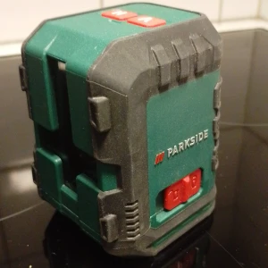 PKLL 10 B4 Grön Cross Line Laser - Parkside PKLL 10 B4 grön korslaser för exakt nivellering. Robust design med gummiskydd, tydliga H/V-knappar och synliga etiketter. Används med 2x1,5V AA-batterier. Mindre yttre damm men i gott skick, perfekt för bygg- och renoveringsprojekt.