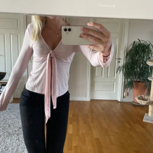 Ljusrosa omlottopp med knyt - Säljer en ljusrosa omlottopp med långa ärmar och djup v-ringning. Toppen har en snygg knytning framtill som ger en feminin touch och är tillverkad i ett mjukt, tunt material. Perfekt att styla med jeans eller kjol för en trendig look. Tröjan är lite nopprig men annars i fint skick!💗