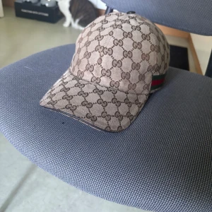 Äkta Gucci keps - Snygg keps från Gucci i beige och brunt med klassiskt GG-monogram över hela kepsen. På sidan finns den ikoniska gröna och röda randen. Kepsen är tillverkad i bomull och har justerbar rem baktill. Perfekt för dig som vill sticka ut med en lyxig accessoar. Lite solblekt vilket knappt syns speciellt när färgen på kepsen är beige. 