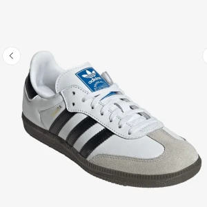 Adidas Samba vita och svarta sneakers - Klassiska Adidas Samba sneakers i vitt skinn med svarta ränder och grå mockadetaljer vid tån. Skon har snörning, platt sula och blå Adidas-logga på plösen. 