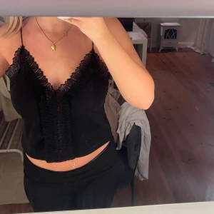 Svart linnetopp med spetsdetaljer - Säljer en svart linnetopp med tunna axelband och snygga spetsdetaljer längs v-ringningen och framtill. Toppen är croppad och har en feminin look som passar perfekt till jeans eller kjol. Materialet känns mjukt och lätt mot huden.