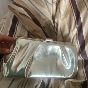 Guldig kuvertväska med kedja - Snygg kuvertväska i metallic guld från NA-KD. Väskan har en stilren kedjerem i guld, hård ram och klassisk låsdetalj upptill. Tillverkad i syntetmaterial med glansig finish. Perfekt accessoar för att lyfta din look. Finns ett kedja som man kan ta fram o lägga i kuvertväskan igen!! Perfekt för bal och annat