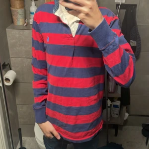 Randig pikétröja från Polo Ralph Lauren - Säljer en klassisk randig pikétröja från Polo Ralph Lauren i slim fit. Tröjan har breda röda och mörkblå ränder, lång ärm och vit krage. Ikoniska blå loggan broderad på bröstet. Perfekt för dig som gillar preppy stil den är m och jag är 177 någon mer skicka då