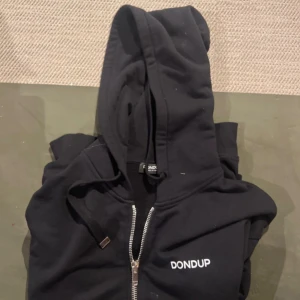 Svart hoodie med dragkedja från Dondup - Svart hoodie från Dondup med vit logga på bröstet, dragkedja framtill och snörning i huvan. Tillverkad i mjuk bomull och har klassisk passform. Perfekt för en chill och stilren look.