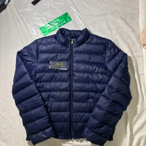Mörkblå dunjacka Polo Ralph Lauren Xs/xl barn - Snygg mörkblå dunjacka från Polo Ralph Lauren i storlek XL barn men passar xs. Jackan är quiltad med horisontella sömmar, har dragkedja framtill och en klassisk ståkrage. Perfekt för kyliga dagar och ger en clean och sportig look.
