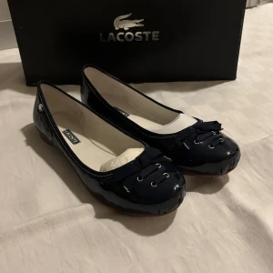 Mörkblå ballerinaskor från Lacoste - Sötaste ballerinaskorna från Lacoste med knytdetaljer framtill och logga på sidan. Använda ett fåtal gånger men ser helt nya ut. Endast sulan har tecken på användning (se bild). Säljes med originalkartongen. Nypris: 899 kr