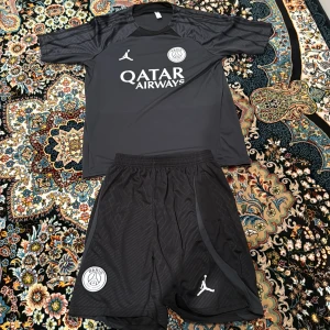 PSG x Jordan svart fotbollströja - Svart PSG fotbollströja med matchande shorts, båda med Jordan-logga och Paris Saint-Germain-emblem. Tröjan har korta ärmar och tryckt Qatar Airways-sponsor på bröstet. Tillverkad i lätt och ventilerande material, perfekt för träning eller match.