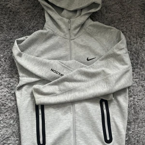 Grå Nike zip up Nocta - Snygg ljusgrå hoodie från NOCTA x Nike med hel dragkedja, svarta detaljer och två fickor med dragkedja. Tröjan har huva, NOCTA-tryck på ärmen och Nike-logga på bröstet. Perfekt för dig som gillar streetwear och sportig stil. Helt ny och oanvänd i storlek M. Byten funkar