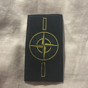 Stone Island patch - Säljer en klassisk svart patch från Stone Island med broderad kompasslogga i gult och grönt. Perfekt att fästa på jacka eller tröja för att ge din outfit en exklusiv och streetinspirerad touch.