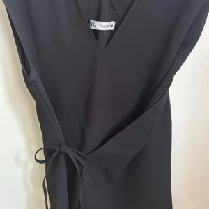 Svart omlotttopp Zara - Stilren svart omlotttopp från Zara med v-ringning och knyt i sidan. Ärmlös modell som ger en clean och modern look. Perfekt för dig som gillar minimalistisk stil och vill ha något enkelt men snyggt.