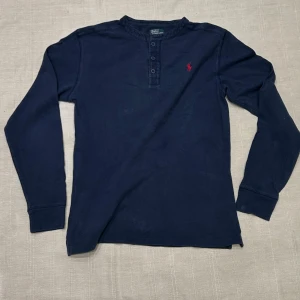 Mörkblå långärmad tröja Polo Ralph lauren - Mörkblå långärmad tröja från Polo Ralph Lauren med rund halsringning och knappslå framtill. Tröjan har en röd broderad logga på bröstet och är tillverkad i mjuk bomull. Perfekt för en clean och sportig stil.