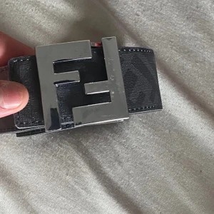 Svart Fendi bälte med FF-spänne - Säljer ett svart bälte från Fendi med klassiskt FF-mönster längs hela remmen och ett stort, blankt metallspänne i silver med Fendi-loggan. Bältet är gjort i skinn och har en stilren, lyxig vibe som sticker ut.