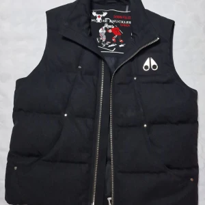Svart dunväst från Moose Knuckles - Svart dunväst från Moose Knuckles med silvrig logga på bröstet och grov dragkedja. Västen har quiltad design och innerfoder med broderat hockeymotiv. Perfekt för lager-på-lager och snygg till streetwear. Ny pris ca 7000, mitt pris 3000 och kvitto medföljer :)