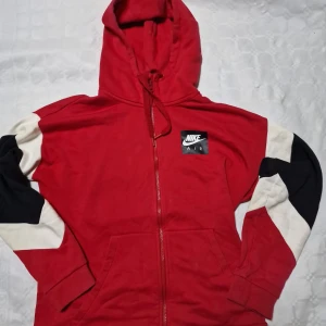 Röd Nike Air hoodie med dragkedja - Säljer en röd hoodie från Nike Air med svart och vit detalj på ärmarna. Tröjan har huva med snörning, hel dragkedja och fickor framtill. Perfekt för dig som gillar sportig stil och streetwear.