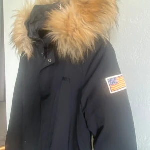 Svart parkas med fuskpäls från Svea - Svart parkas från Svea med avtagbar fuskpäls på huvan och amerikansk flagga på ärmen. Jackan har dragkedja och knappar framtill, två fickor med lock och broderad Svea-logga på bröstet. Yttertyg i bomull och nylon, fodrad med polyester.