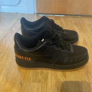 Nike Air Force 1 Gore-Tex  - Svarta Nike Air Force 1 Gore-Tex sneakers med orange broderade detaljer och loggor. Skorna har en klassisk låg siluett, snörning och är tillverkade i en mix av skinn och syntetmaterial för extra hållbarhet och vattentålighet. Perfekta för dig som vill ha stil och funktion.
