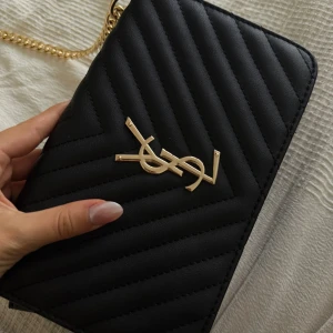 Svart quiltad YSL axelväska i skinn - ❣️Obs!! Reser från den 11/9-12/10 och kommer ej ha möjlighet att skicka något då, så passa på 🤗priset kan ofta diskuteras ❣️                                   Säljer en svart quiltad axelväska från YSL med ikonisk guldlogga framtill. Väskan har en snygg kedjerem i guld och insida i rött. Perfekt storlek för det viktigaste och ger en lyxig vibe till din outfit. Klassisk design med elegant form och detaljer.