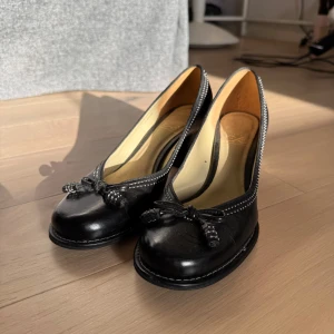 Svarta pumps från Clarks med rosett - Snygga svarta pumps från Clarks i skinn med rund tå och dekorativ rosett framtill. Skorna har låg klack och kontrastsömmar längs kanten för en klassisk look. Perfekta för dig som gillar tidlös stil med en twist.