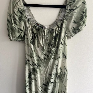 Grön mönstrad kortklänning Hostess - Superfin kortklänning från Hostess i ljusgrönt och mörk gröna mönster. Klänningen har puffärmar, knytning vid bysten och en söt, rynkad urringning. Perfekt för dig som gillar en trendig och avslappnad stil.