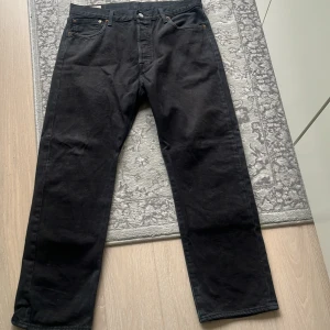 Svarta Levi's 501 jeans 36/30 - Klassiska svarta Levi's 501 jeans i rak modell. Jeansen är tillverkade i kraftigt denimtyg och har fem fickor, knappgylf och läderpatch bak i midjan. Perfekta för dig som gillar en tidlös och clean look.