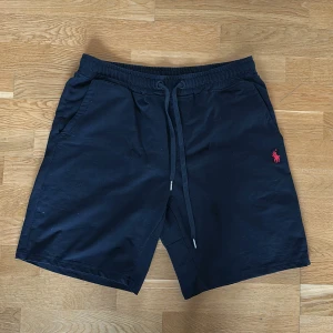 Mörkblå shorts från Polo Ralph Lauren - Snygga mörkblå shorts från Polo Ralph Lauren med klassisk röd logga broderad på framsidan. Modellen har elastisk midja med snörning, två sidofickor och en bakficka. Perfekta för chill dagar och sommarhäng.