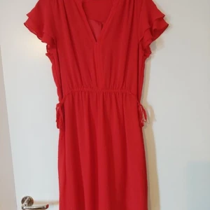 Röd kortklänning med volangärmar stl M - Snygg röd kortklänning med v-ringning och volangärmar. Klänningen har resår i midjan med knytband i sidorna för en skön passform. Lätt och luftig modell, perfekt för varma dagar. Stilren design utan mönster och med mjukt, tunt tyg.