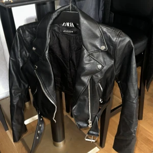 Svart skinnjacka från Zara, S - Säljer en svart skinnjacka från Zara i storlek S. Jackan har klassisk bikerstil med silvriga dragkedjor, knappar och bälte i midjan. Snygg krage och detaljerade sömmar ger en cool vibe. Perfekt för dig som vill ha en edgy look. 