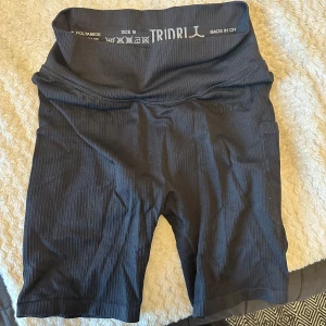 Svarta ribbade cykelshorts TRIDRI - Svarta ribbade cykelshorts från TRIDRI med hög midja och tight passform. Perfekta för träning eller chill. Materialet är stretchigt och andas bra. Snyggt diskret mönster och bekväm midja.