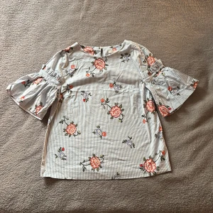Blommig randig blus med volangärm från H&M - Supersöt blus från H&M i ljusblått och vitt med tunna ränder och broderade blommor i rosa, rött och lila. Blusen har vida volangärmar och rak passform. Perfekt för dig som gillar romantiska detaljer och fräscha färger. Aldrig använd. Säljer pga fel storlek. 