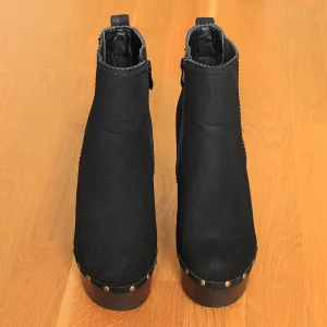 Svarta klackskor - Svarta skor med hög klack och platå. Skorna har snygga guldfärgade nitar längs sulan och dragkedja på insidan. Ovandelen är i mockaliknande material och sulan är i mörkbrunt träl. Perfekta för dig som vill sticka ut lite extra. Är enbart provade och aldrig använda.