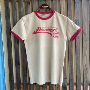 Beige röd ringer t-shirt Düsseldorf  - Retroinspirerad beige ringer t-shirt från Jt's Ultimate med röda kontrastkanter och Düsseldorf-tryck framtill. Klassisk passform och cool vintagekänsla, perfekt för dig som gillar oldschool vibes. Tillverkad i mjuk bomull. Snarare storlek S, nästan XS än M. 