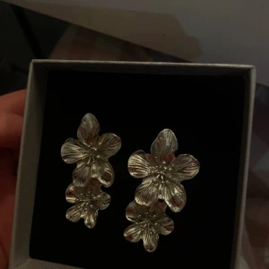 Silvriga blommiga örhängen - Supersnygga örhängen i silverfärg med dubbla blommor på varje örhänge. De har en detaljerad, glansig yta och ger en riktigt cool statement-look. Perfekta för dig som vill sticka ut med ett unikt smycke med blommig design.