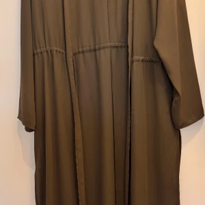 Olivgrön lång kimono - Säljer en olivgrön lång kimono med vida ärmar och avslappnad passform. Materialet känns mjukt och faller snyggt. Perfekt att slänga över en outfit för en cool lager-på-lager-look.