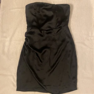Svart axelbandslös klänning Bik Bok XS - Helt ny svart axelbandslös kortklänning från Bik Bok i glansigt satintyg. Klänningen har en figurnära passform och dold dragkedja i ryggen. Perfekt för dig som vill ha en stilren och elegant look.