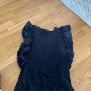 Svart volangblus med ärmlös design - Säljer en svart blus med volangdetaljer längs axlarna. Toppen är ärmlös och har en rund halsringning. 