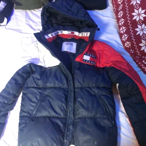 Tommy Jeans pufferjacka XS passar bra på S också - Snygg pufferjacka från Tommy Jeans i färgblockad design med marinblå, röd och vit. Jackan har huva, dragkedja framtill och två fickor med dragkedja. Ikonisk Tommy Jeans-logga på bröstet och detaljer på insidan av kragen. Ny pris 1800kr