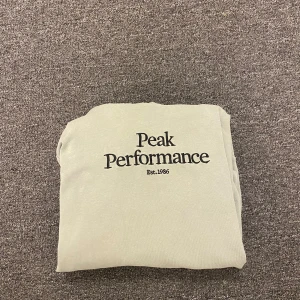 Ljusgrön hoodie från Peak Performance - Snygg ljusgrön hoodie från Peak Performance med svart logga på bröstet. Klassisk känguruficka framtill, ribbade muddar och huva. Perfekt för chill dagar eller när du vill ha en enkel och stilren look.