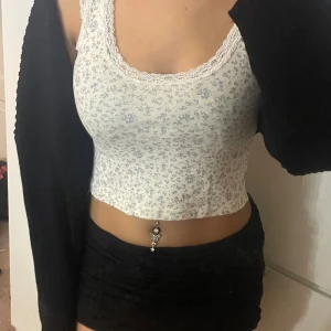 Blommig croppad linnetopp Brandy Melville - Supersöt croppad linnetopp från Brandy Melville i vit med ljusblått blommigt mönster. Toppen har spetsdetaljer runt halsringningen och är ärmlös med rundad form. Perfekt för sommarens alla dagar, mjuk och stretchig bomull. ”No size” men passar S💕💕pris kan diskuteras